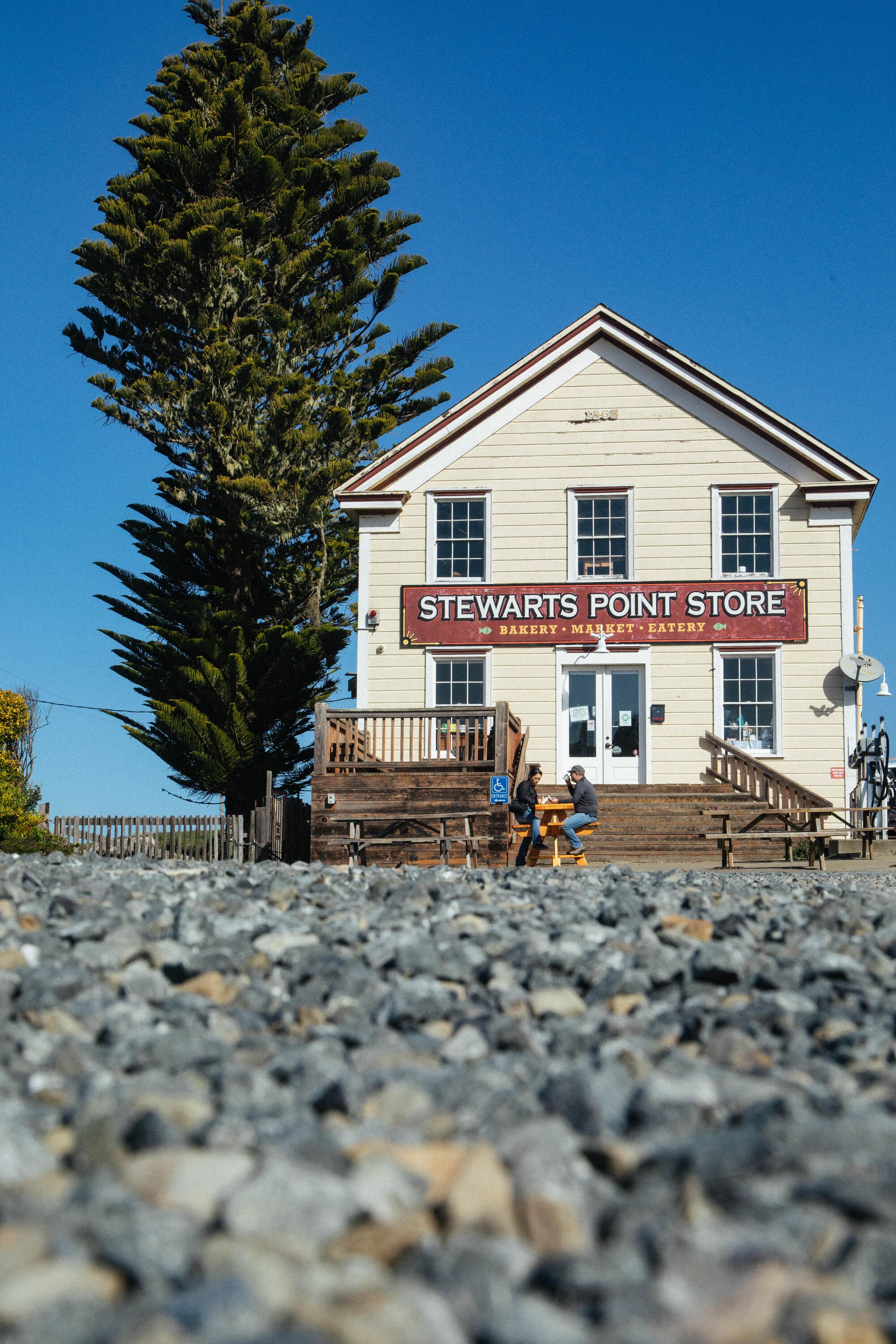 A cream-colored wooden building, with a sign that says Stewarts Point Store, next to a large tree.