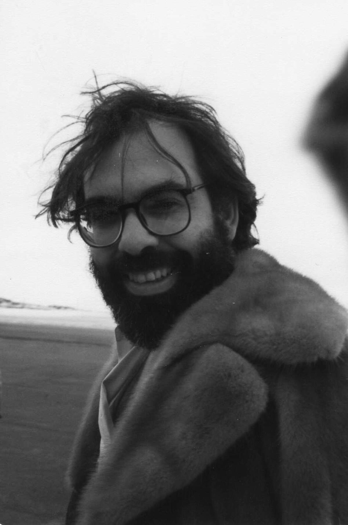 Francis Ford Coppola, a smiling man with an unruly beard and hair, stands on a beach wearing a fur coat.