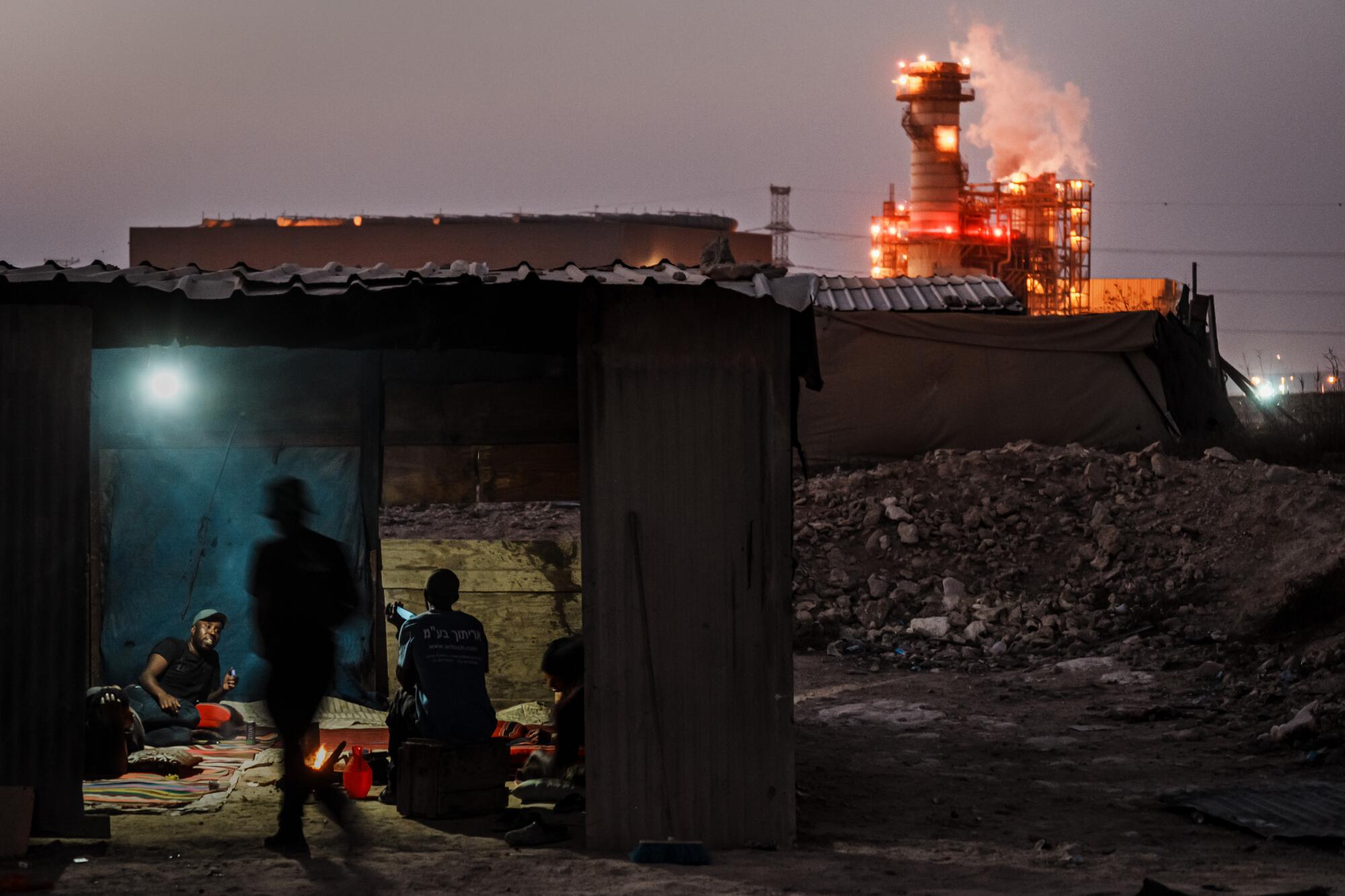  People shelter under a structure with a roof and open sides. A power plant glows and spews smoke in the distance 