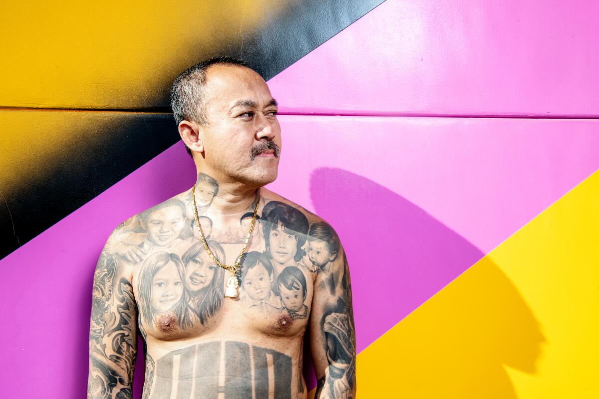 A man with tattoos on his torso and arms stands against a colorful wall