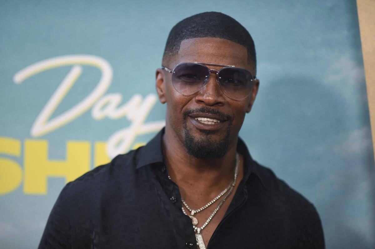 Jamie Foxx in aviator sunglasses , black dress shirt and necklaces smiling in front of a light blue backdrop