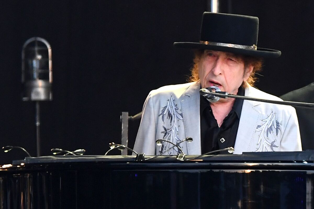 A man in a black hat sings into a microphone while seated at a piano.