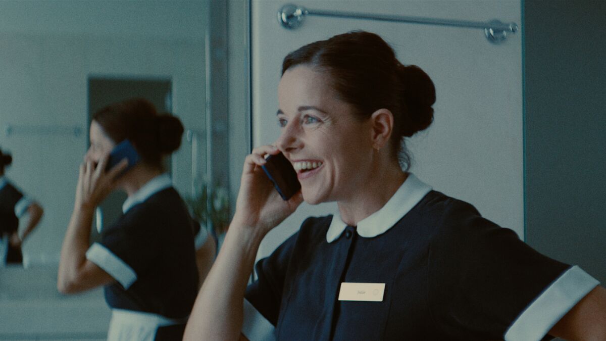 A woman in a maid's uniform, speaking on a cellphone and smiling, with her reflection in a mirror.