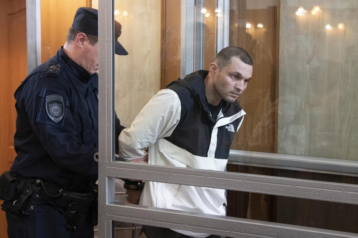 A guard escorts a handcuffed man into a glass cage in a Russian courtroom.