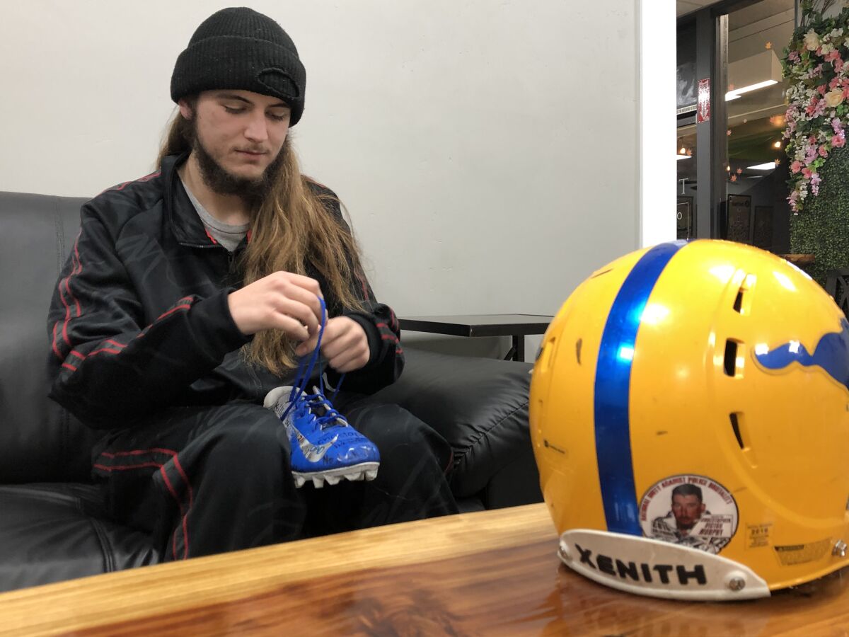 Gabe Murphy ties the laces on one of his football cleats, and a helmet on the table has a sticker of his late father