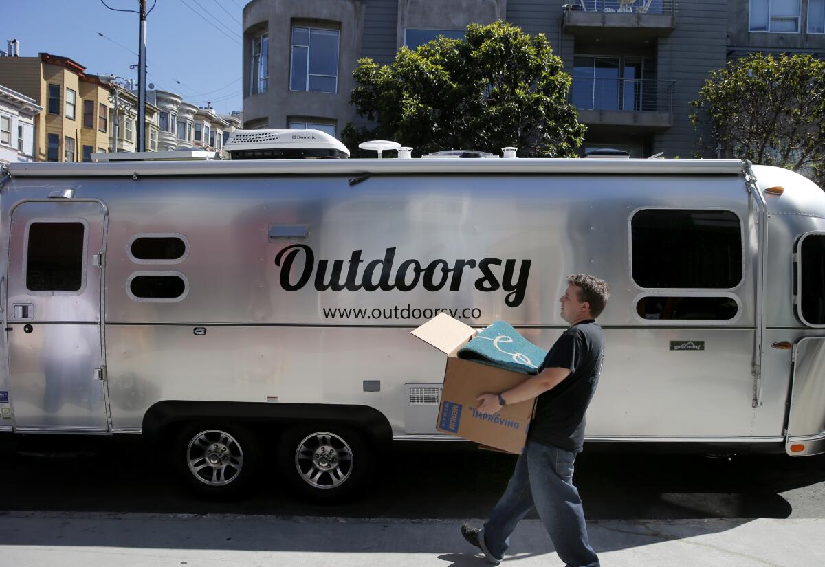A man carries a full cardboard box to an Airstream with the word "Outdoorsy" on it.