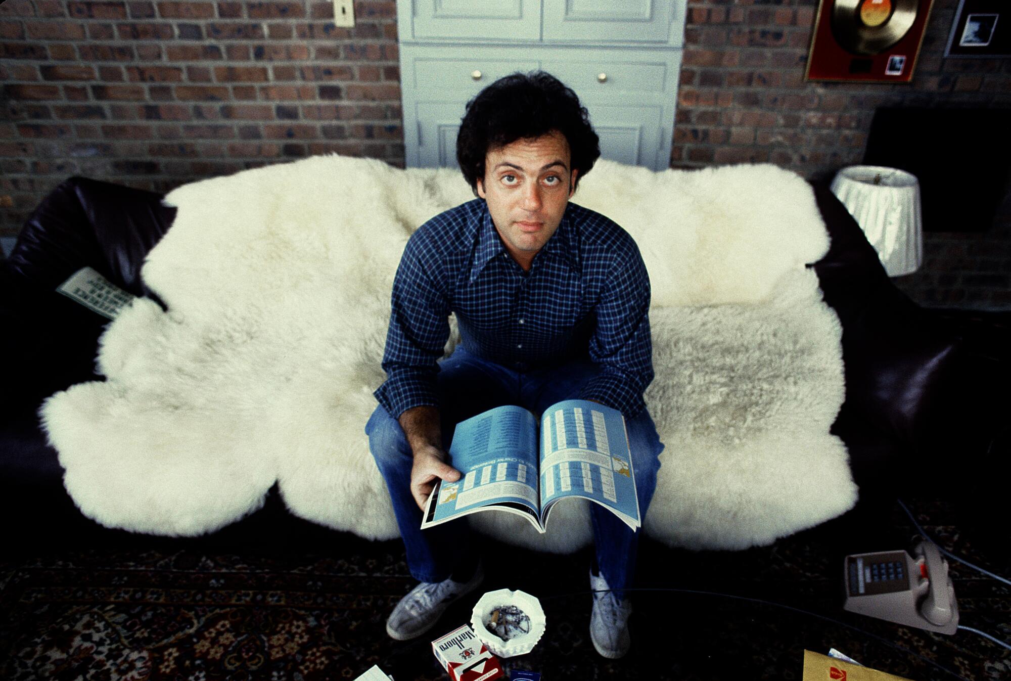 A young man sits on a sheepskin-covered sofa holding an open magazine