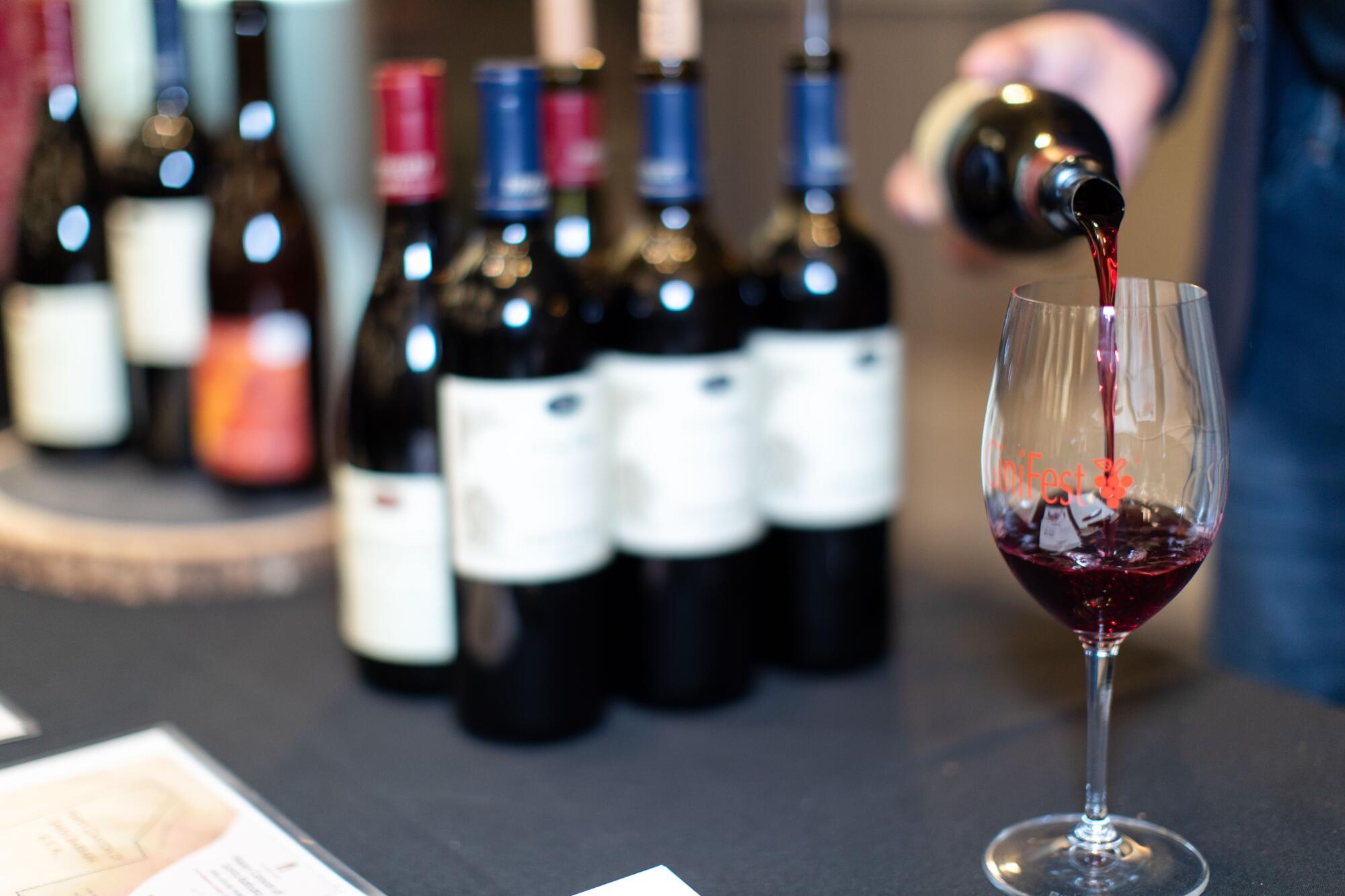 A hand pours wine into a glass on a counter where there are several bottles of wine standing.