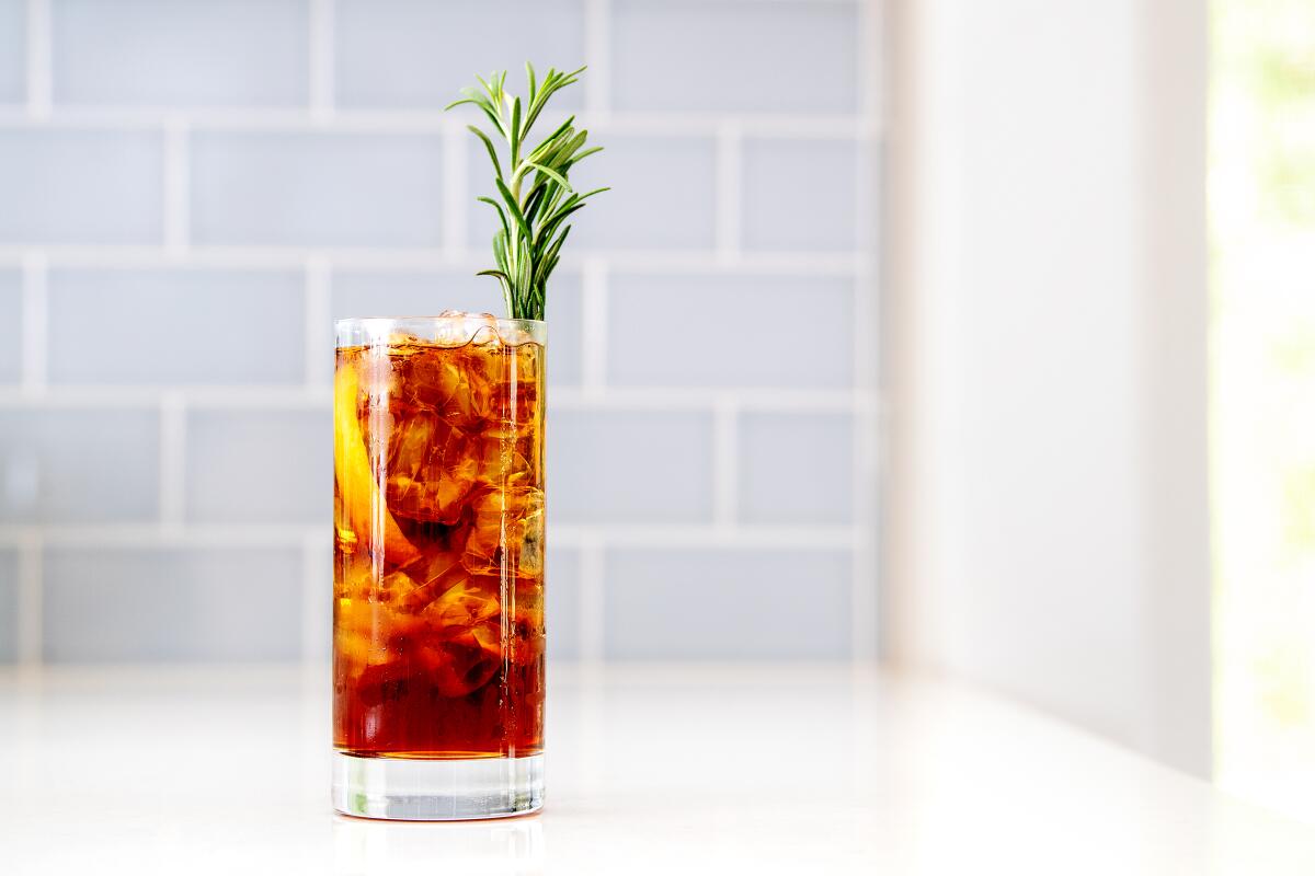 A brown drink in a glass with ice and a lemon and rosemary garnish