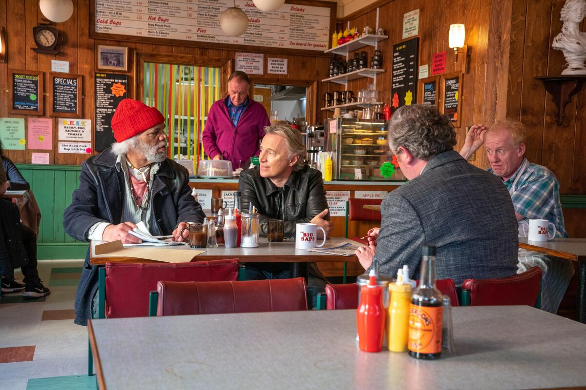 Four men sit at a restaurant table with red chairs. A man is standing in the back behind the counter.