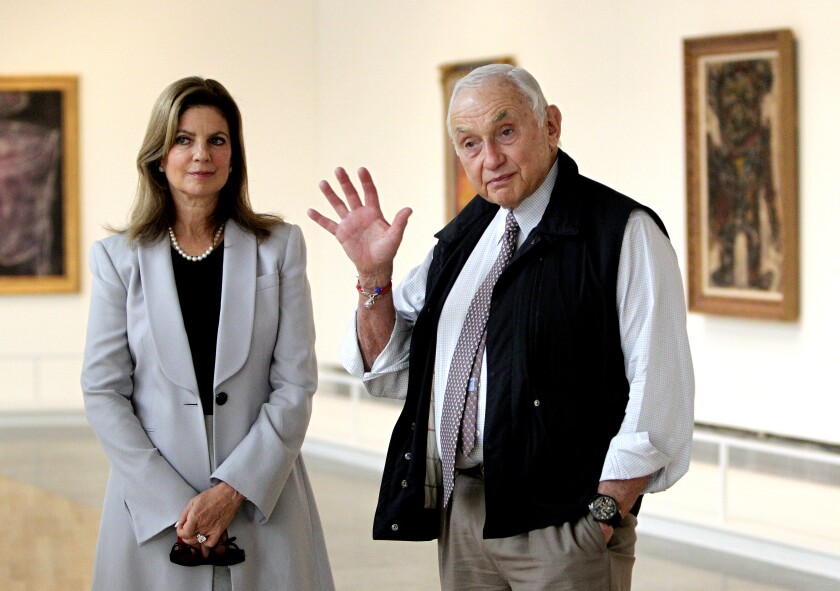 An older man with white hair and his younger wife stand in an art gallery