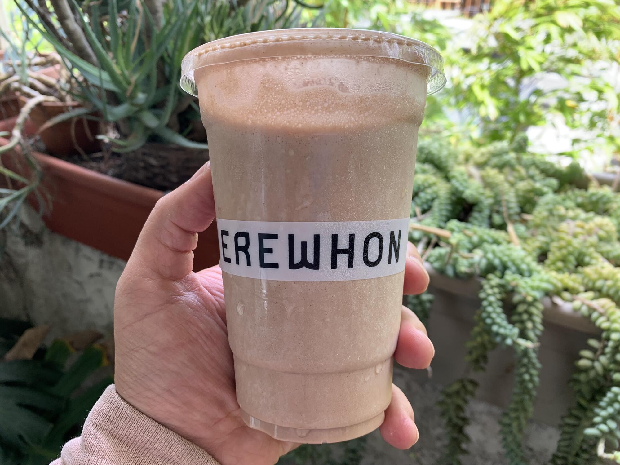 A hand holds a light brown smoothie in a plastic cup that says Erewhon.