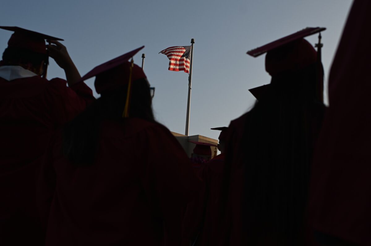 An American flag flies above a building near students at a college graduation ceremony.