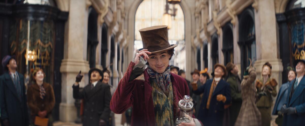 Timothée Chalamet tips his brown top-hat and smirks in front of a crowd of people looking skyward.