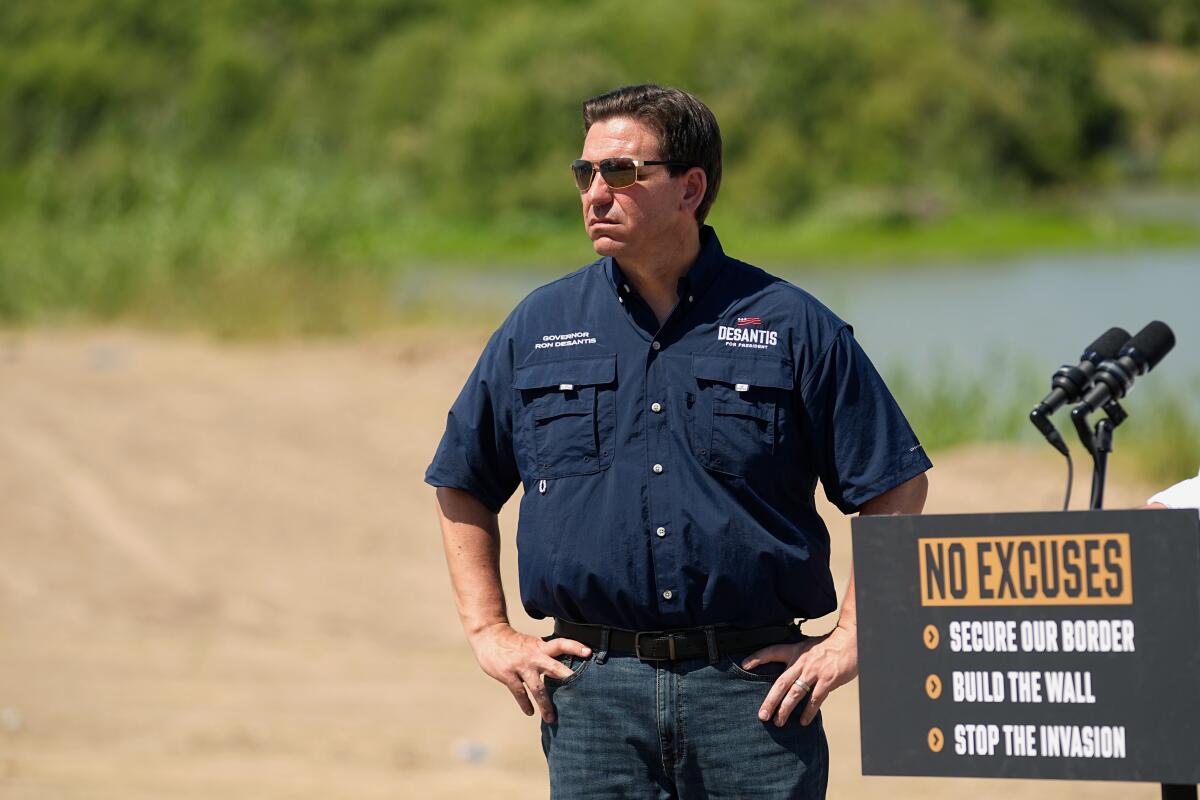 Ron DeSantis, wearing jeans and dark short-sleeved shirt and sunglasses, stands outside in the middle, hands on his hips,