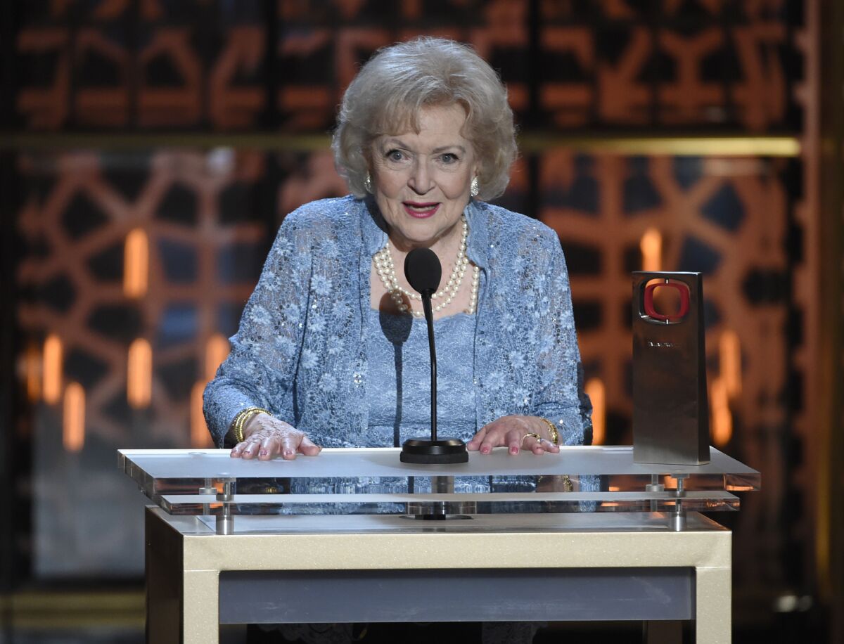 An older woman with white hair in a blue outfit speaks at the microphone at an awards show