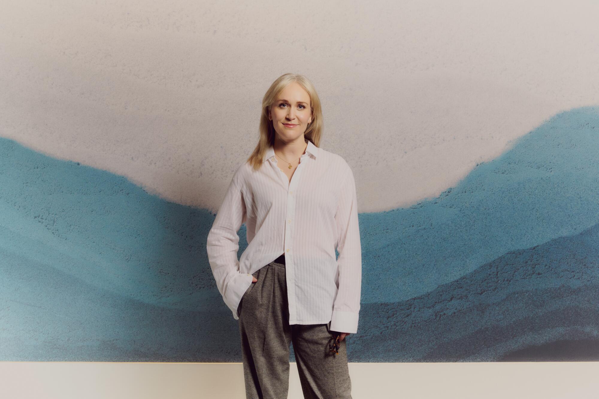 A blond woman, wearing a white button-down shirt and gray trousers, poses with her right hand in her pocket.