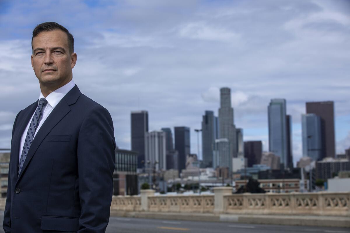 A man wears a white shirt and dark jacket with the L.A. skyline behind him. 