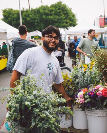 A bearded smiling man carries a bucket of flowers at an outdoor farmers market