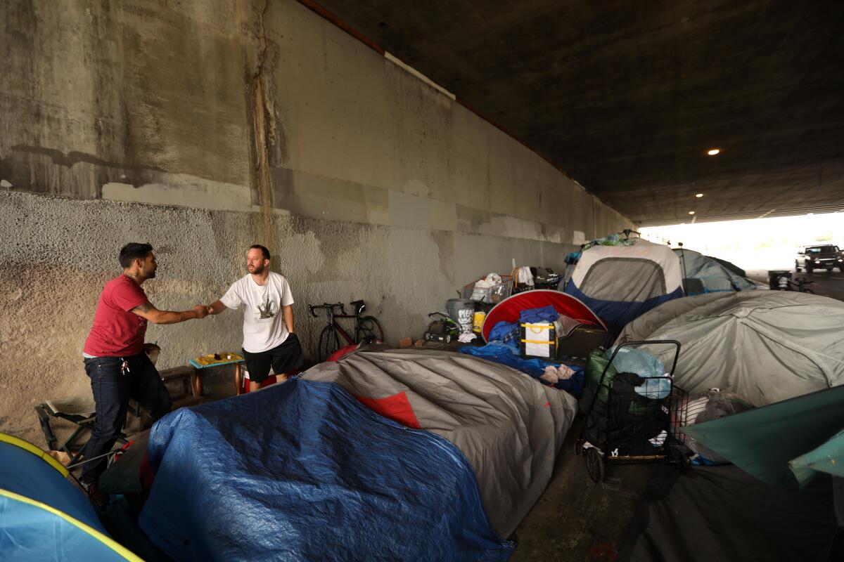 Two men fist-bump amid tents under a freeway overpass
