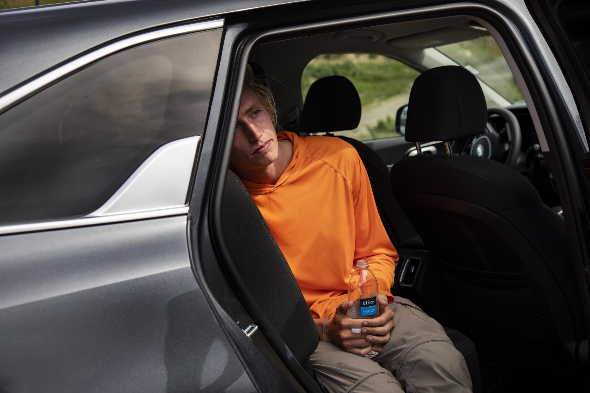 Parell rests with a drink in the back seat of a car with an open door 