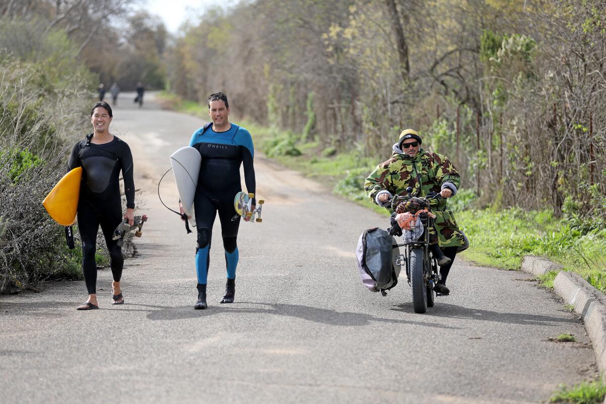 A man on an e-bike passes two surfers walking on a trail. 