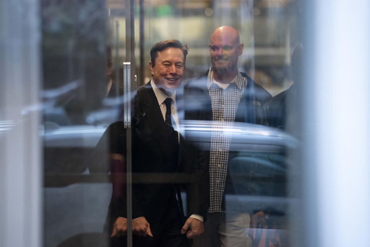 Elon Musk, in a suit and tie, is seen through a glass door.