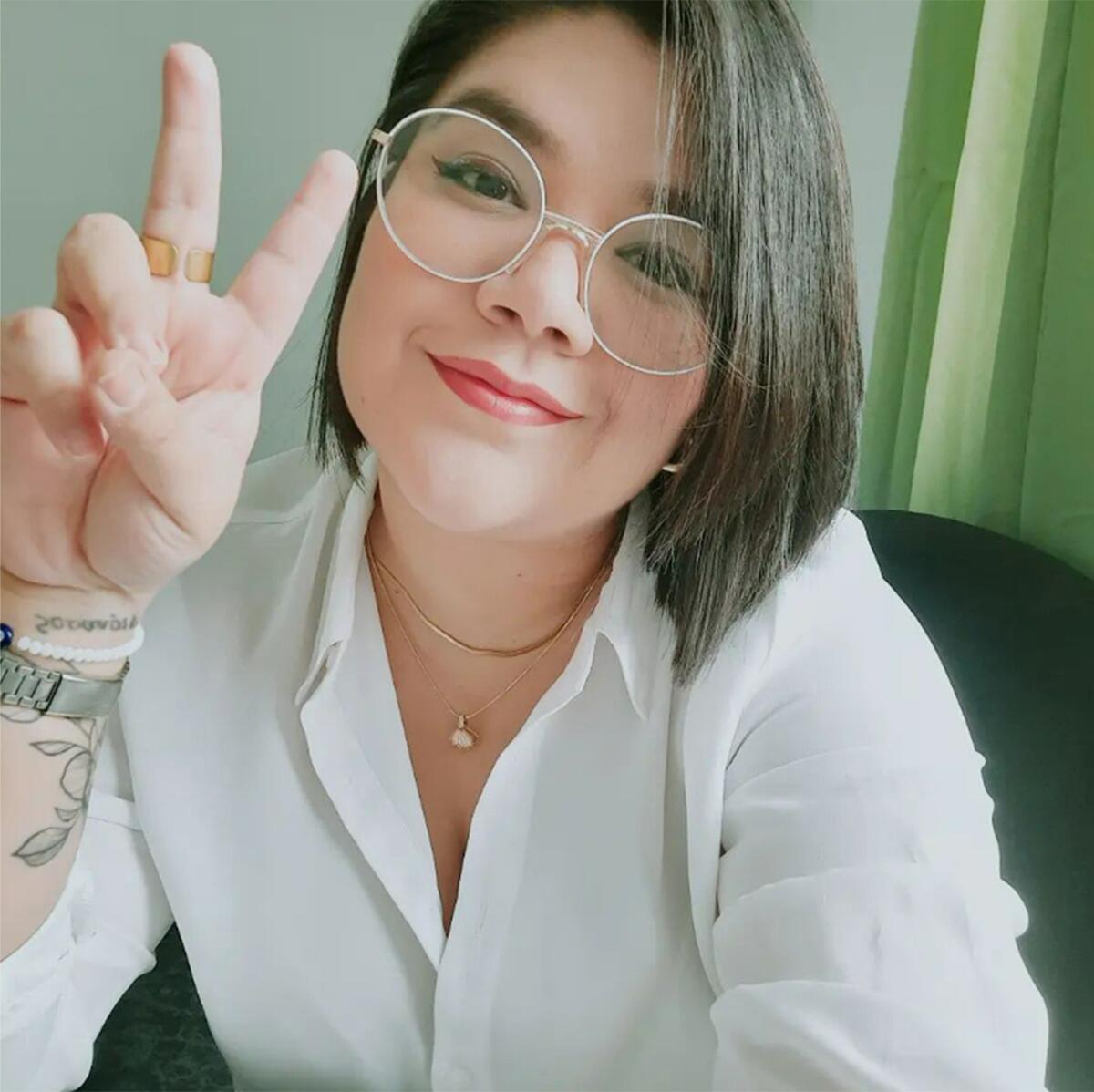 A woman with short dark hair smiles into the camera and flashes the 'V" sign. 