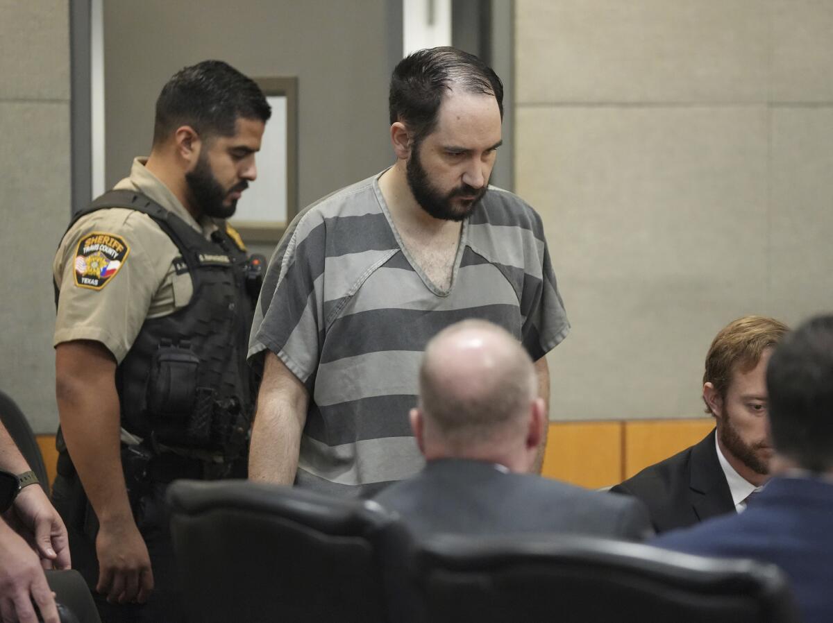 A balding man with a beard and wearing a striped jail top enters court. 