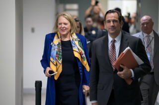 A woman with a blond bob haircut, in a blue coat and colorful yellow scarf, walks behind a man in a suit and striped tie