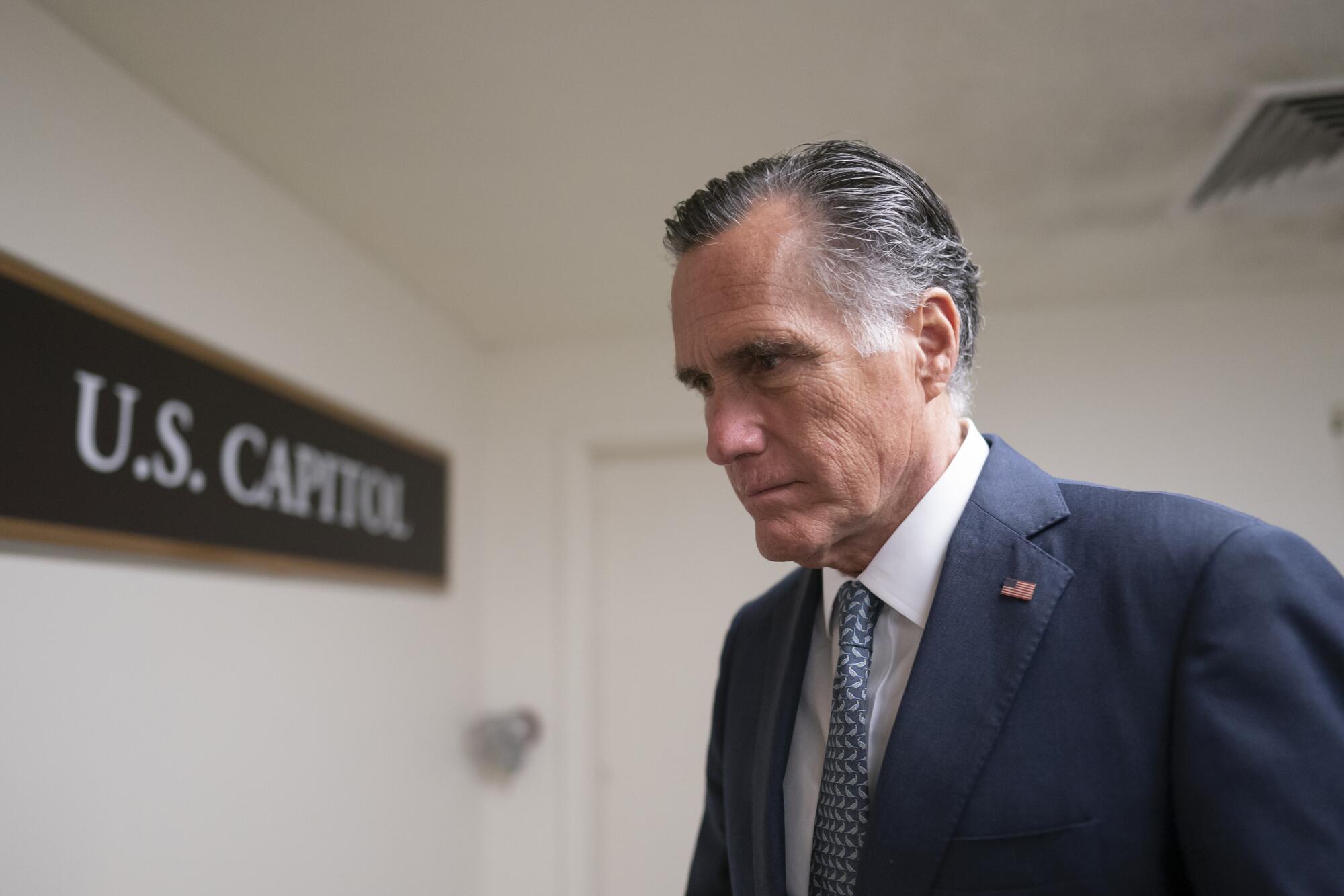 Mitt Romney, wearing a dark suit and white dress shirt, walks past a sign that reads "U.S. Capitol."