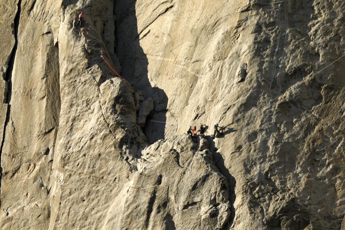 From afar, a few climbers stand on the ledge on a mountainside made of rock