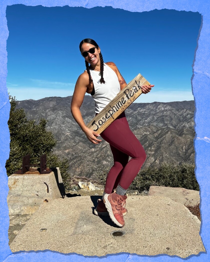 A woman in braids, sunglasses and yoga pants stands atop a rock, smiling and holding a sign that says "Josephine Peak."