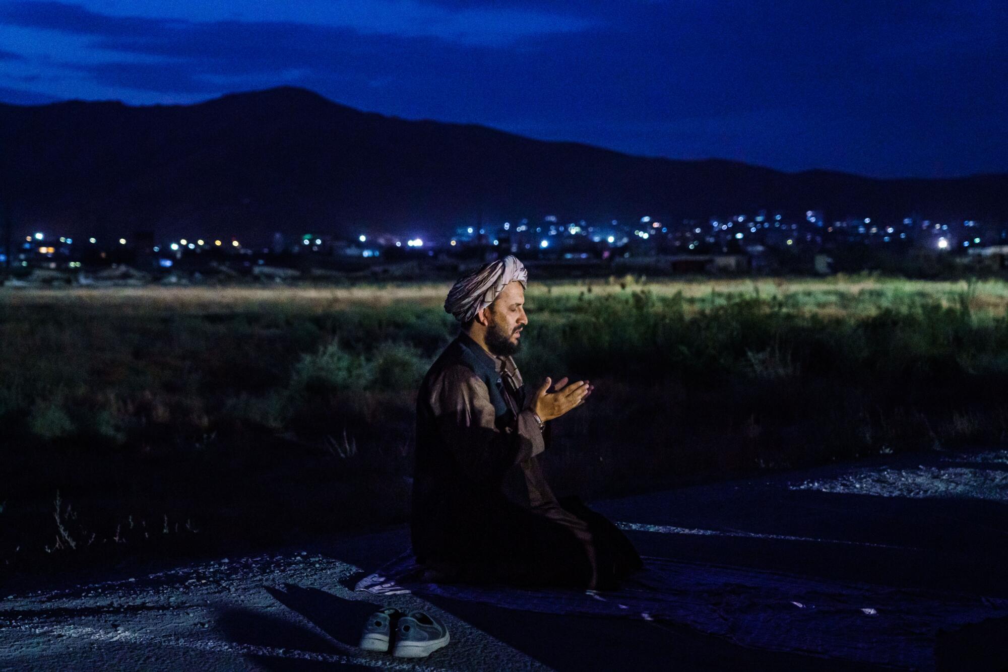 A Taliban fighter kneels in the dark while city lights are in the distance behind him