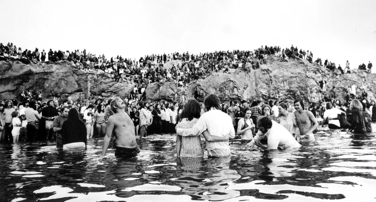 People on a beach watch other people standing, embracing and being baptized in the water.