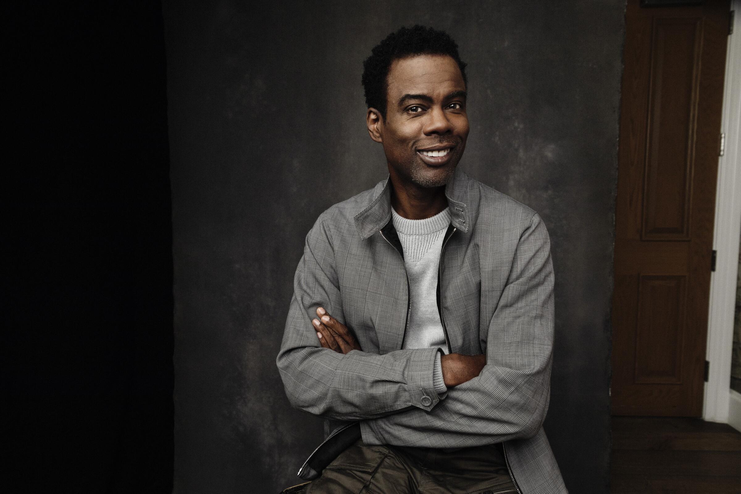 A portrait of Chris Rock, smiling and wearing a jacket with his arms crossed.