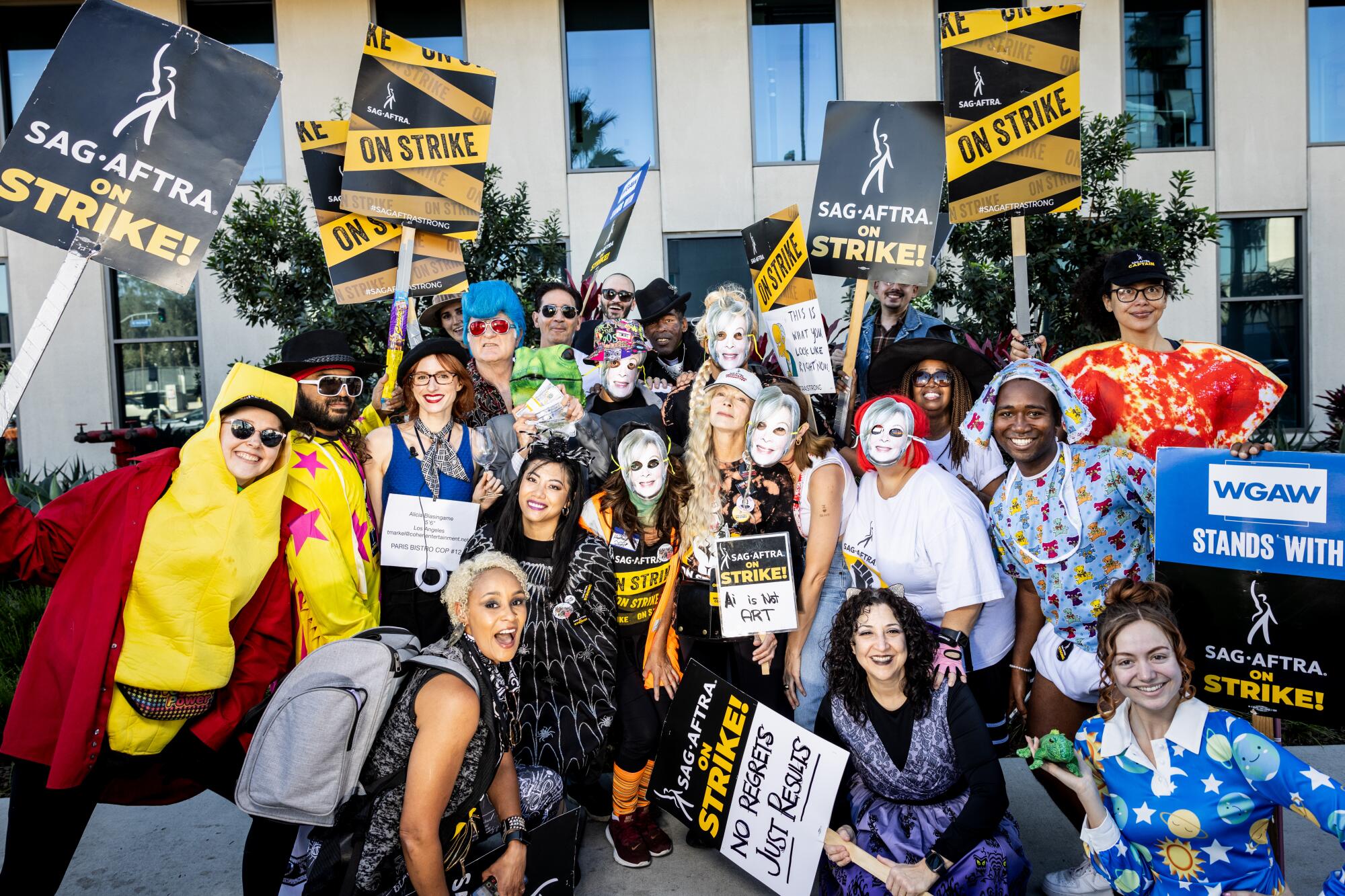 A group photo of actors, most in masks or other costume and many holding picket signs