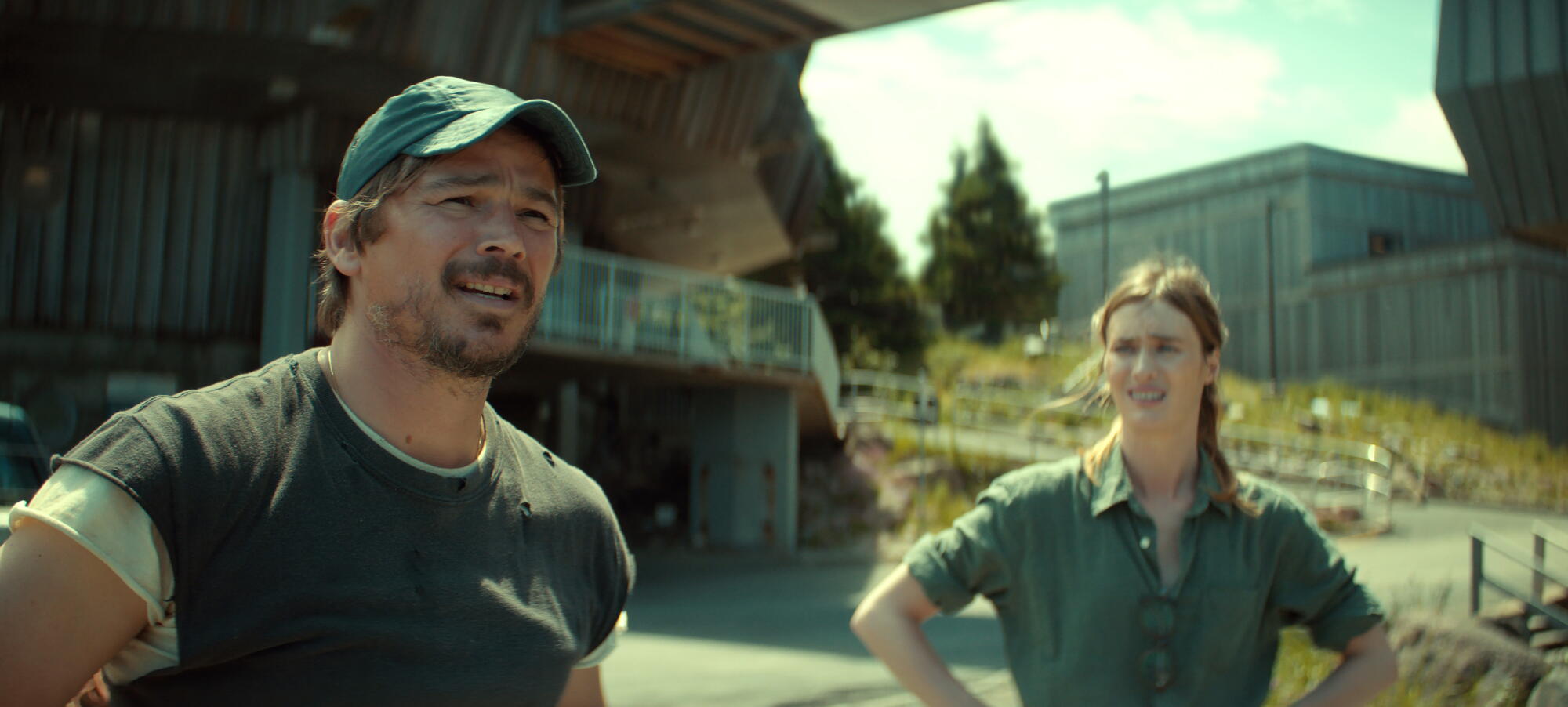 A man in a T-shirt and cap with a concerned expression stands near a woman in a blue shirt outdoors.