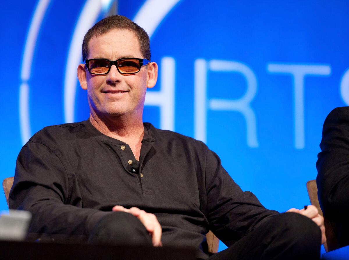 Mike Fleiss, wearing glasses and a black shirt, sits on a stage.