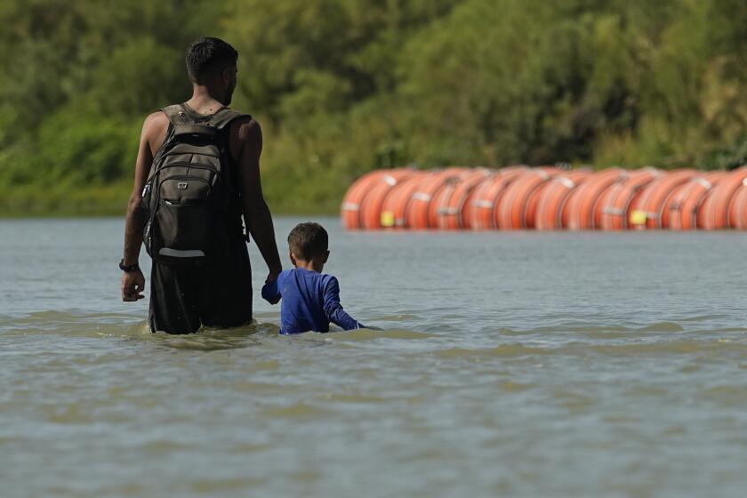 FILE - Migrants walk past large buoys being used as a floating border barrier on the Rio Grande, Aug. 1, 2023, in Eagle Pass, Texas. Texas attorneys asked federal appeals court judges Thursday, Oct. 5, 2023 to let the state keep in place a floating barrier of large concrete-anchored buoys to block migrants from crossing the Rio Grande — a barrier the Biden administration says was illegally deployed without needed federal authorization. (AP Photo/Eric Gay, file)