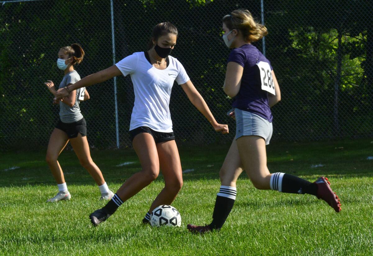 Three high school girls play soccer outdoors in masks.