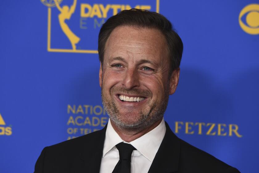A man with dark hair and short facial hair wearing a black suit and tie smiling and posing against a blue and gold backdrop