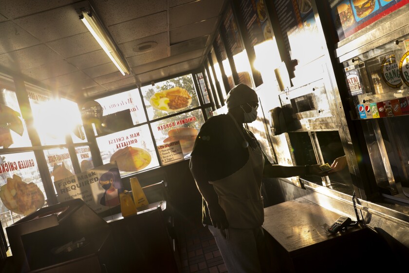 A woman wearing a mask stands inside a takeout restaurant and hands cash through a small window.