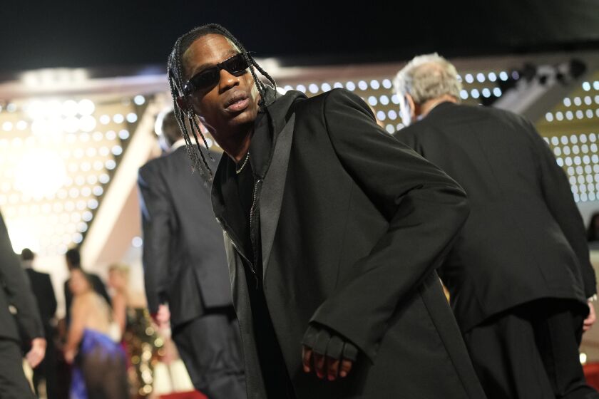 Travis Scott looks over his shoulder in black sunglasses, suit and gloves