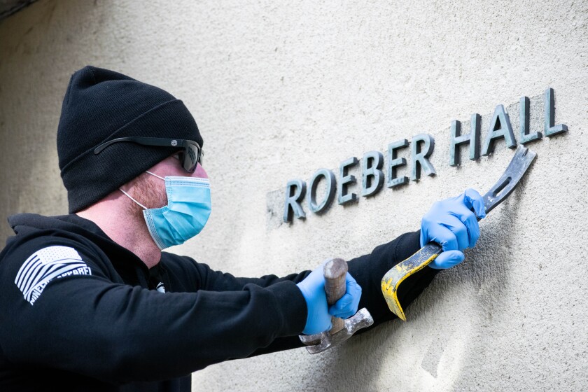 A man uses a hammer and chisel to remove letters spelling "Kroeber Hall" from a wall