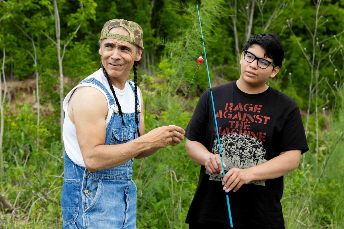A man in a white shirt and overalls stands next to a boy in a Rage Against the Machine T-shirt holding a fishing pole.