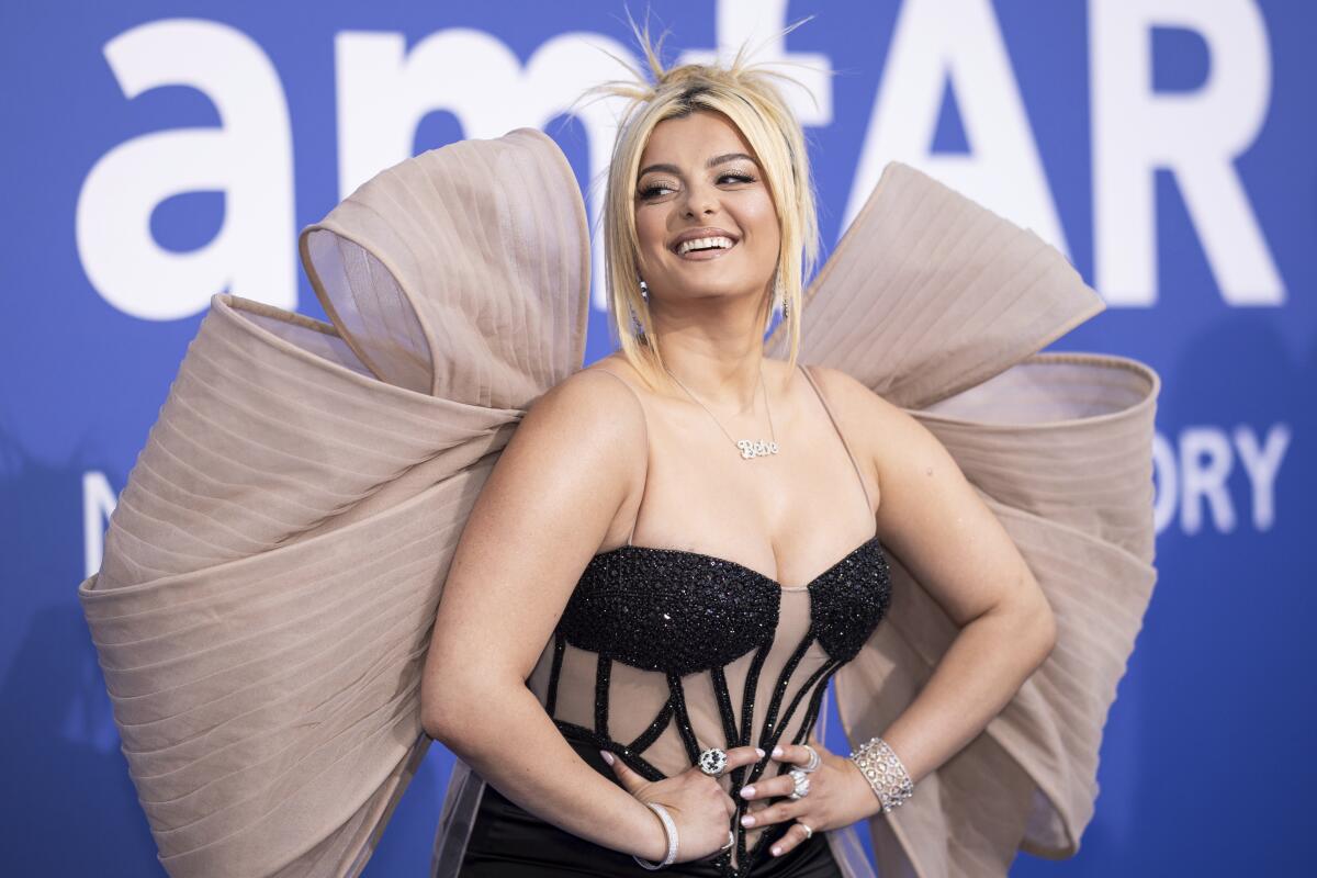 Bebe Rexha with a messy bun, wearing a black and sheer dress with a big bow, poses holding her hands on her waist