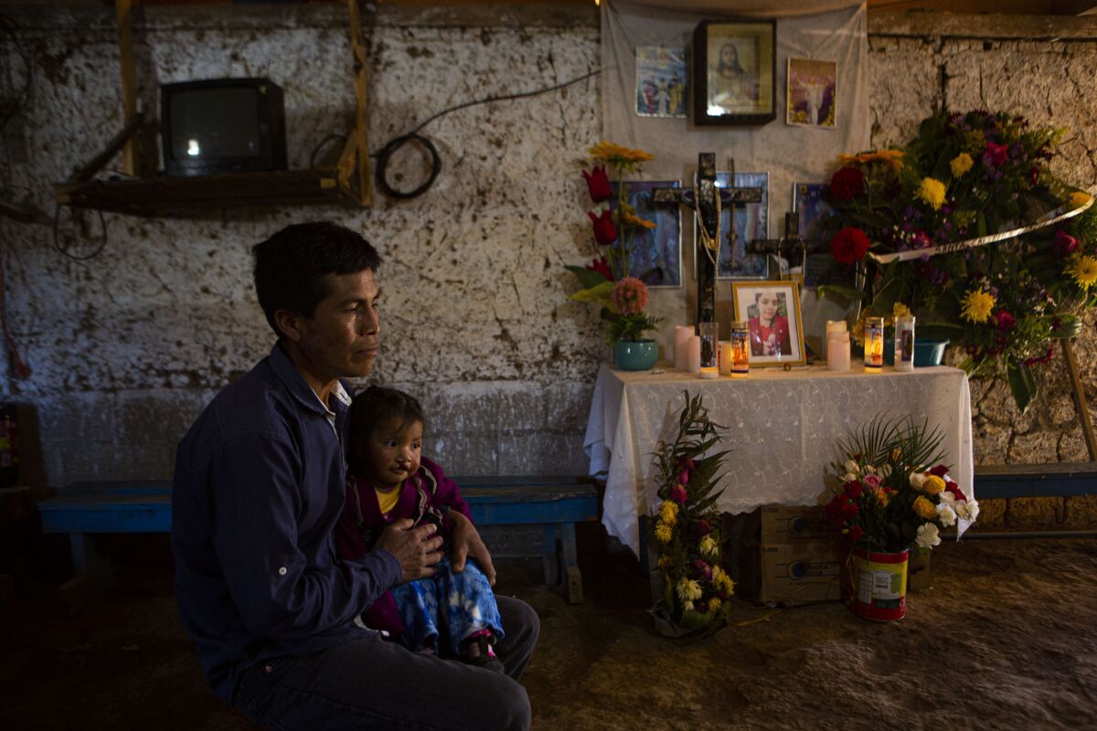 A man sits with his daughter near an altar with candles and flowers and a girl's photograph