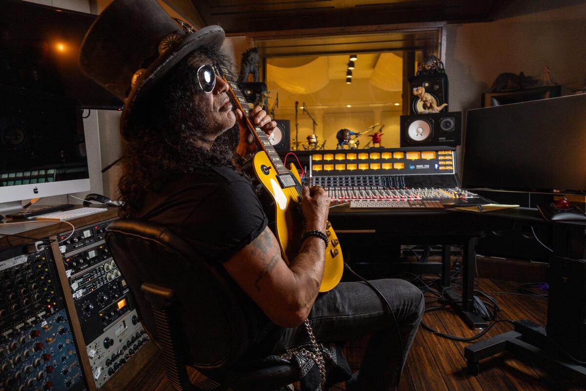 Man in top hat and glasses sits in a recording studio playing guitar 