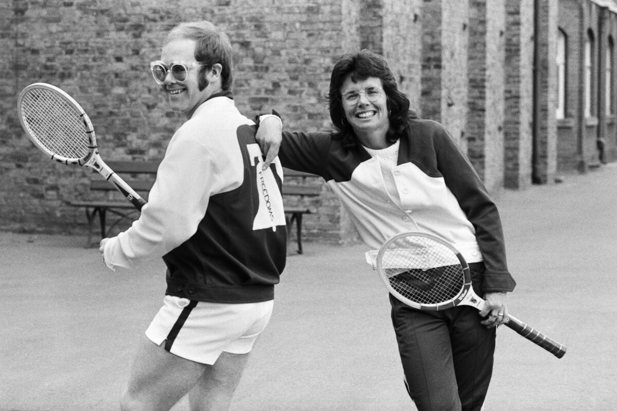 Billie Jean King leans against Elton John's back. Both hold tennis rackets in a black-and-white photo.